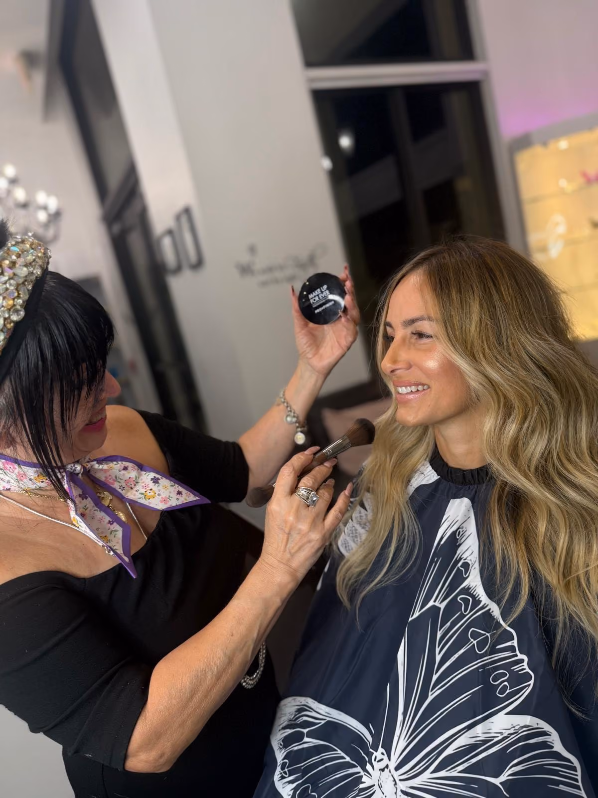 Mimi applying makeup at the chair with a brush, a Make Up For Ever Pro Finish compact in her raised hand.